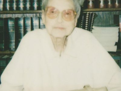 A gray-haired woman with glasses seated in front of a library of books