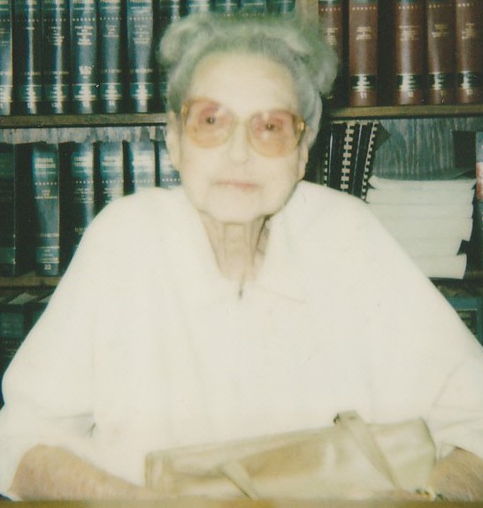 A gray-haired woman with glasses seated in front of a library of books